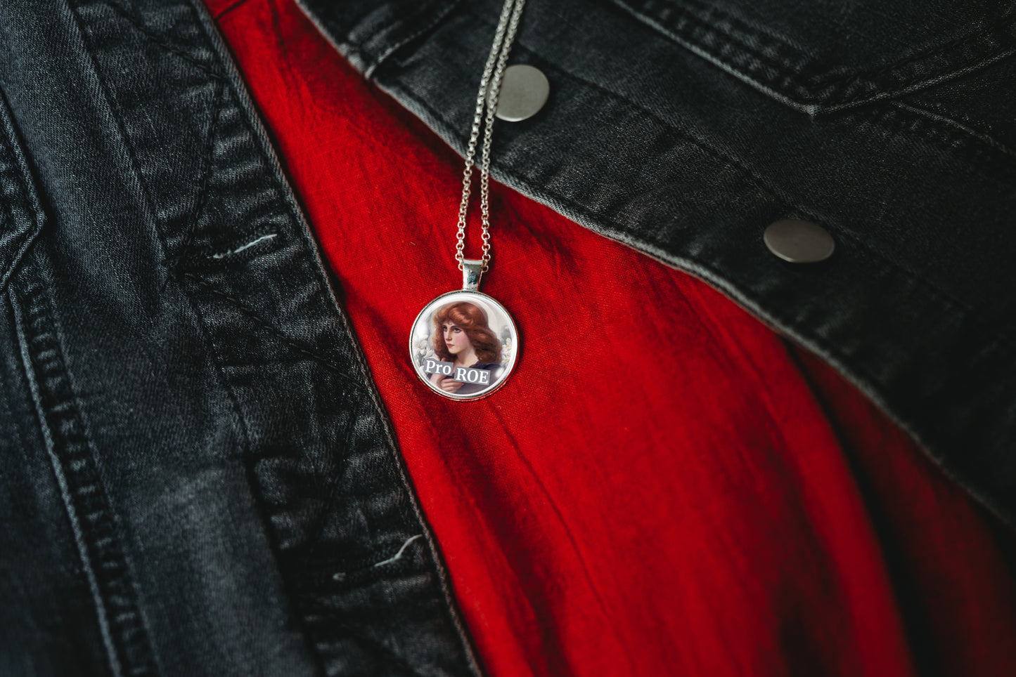 A silver pendant necklace featuring a circular photo print with the text 'Pro ROE' under a woman's face, hanging from a silver chain against a red shirt and black denim jacket.