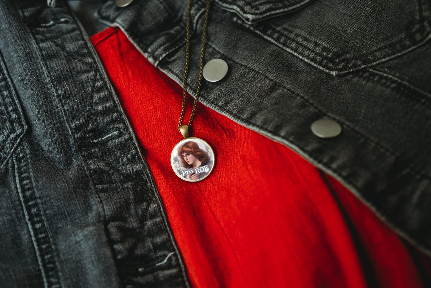 A close-up of a circular pendant necklace with a photo and the text 'Pro ROE' on a bronze chain, styled over a bright red shirt and a dark gray denim jacket.