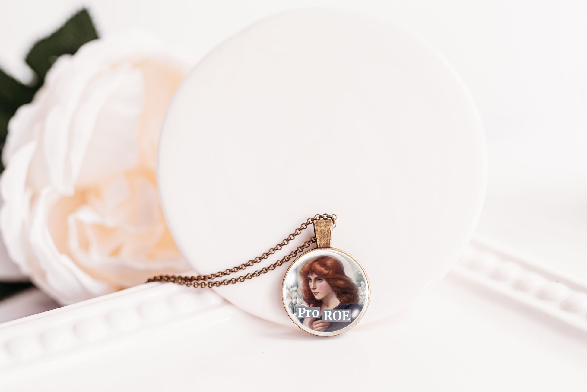 A round pendant necklace featuring a vintage-style portrait of a woman with the text 'PRO ROE' on the front, hanging from a delicate chain and styled on a white surface next to a soft white flower.