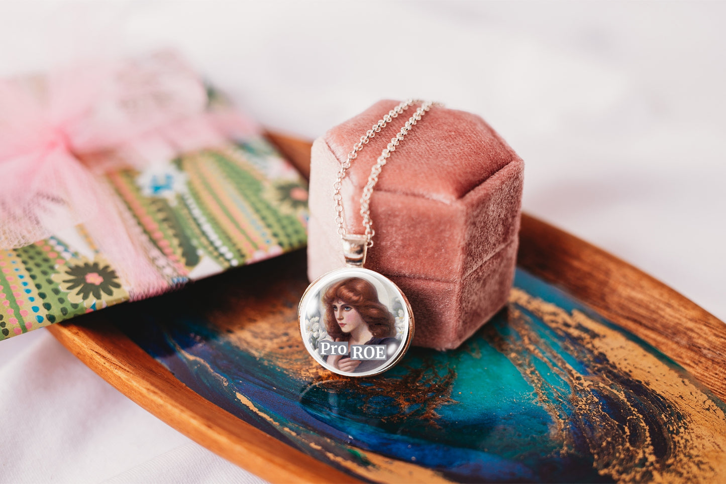 A round pendant necklace with a retro image of a red-haired woman, displayed on a dusty rose velvet jewelry box set on a wooden tray with a colorful design