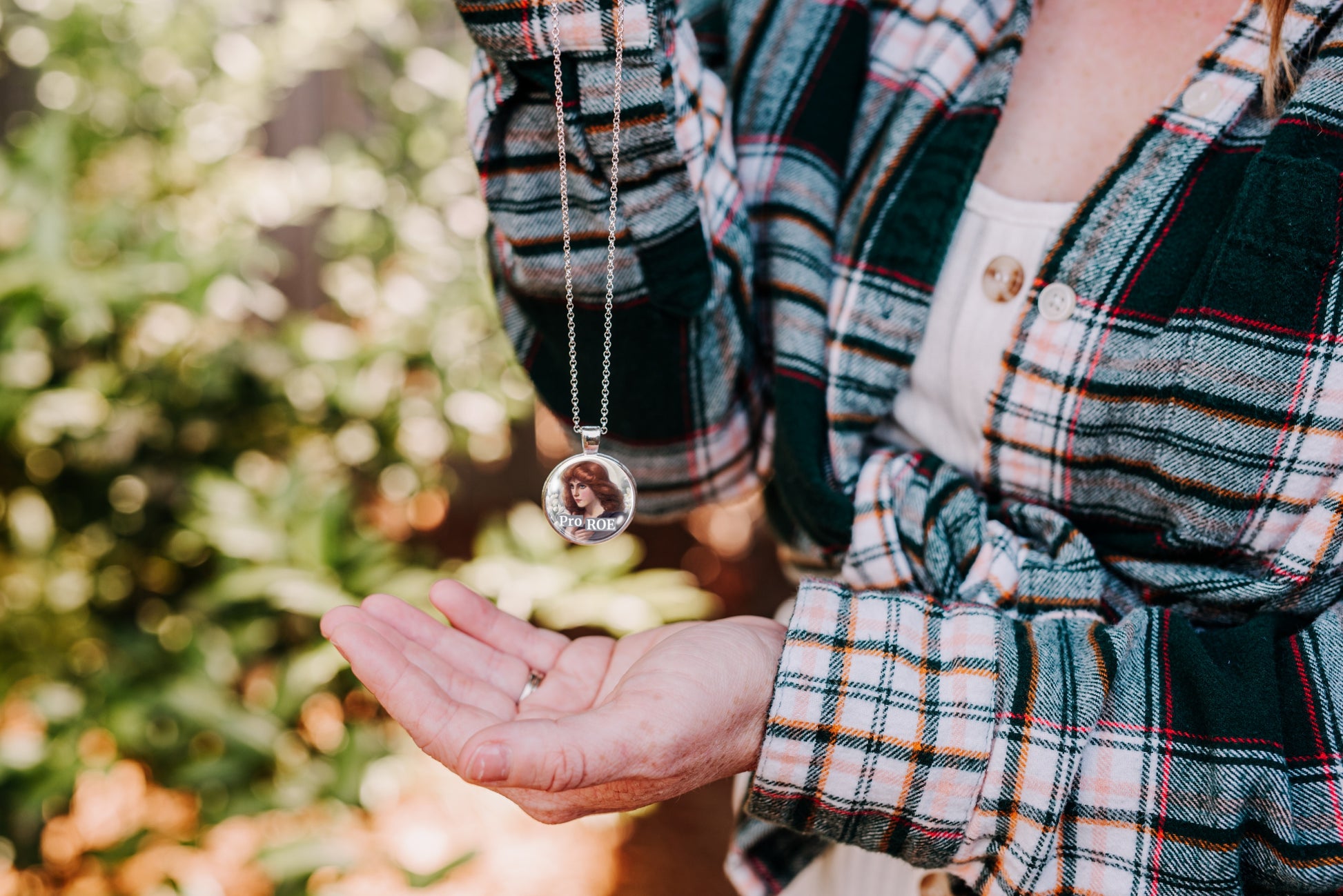 Person wearing a plaid shirt with a necklace featuring a round pendant, set against a blurred natural background.