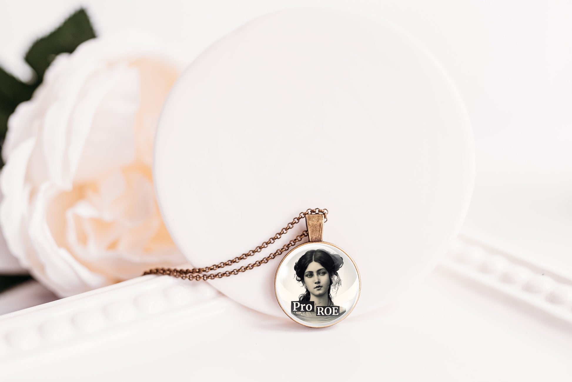 A round pendant necklace with a vintage portrait of a woman and the text 'Pro ROE' on a delicate chain, styled on a white surface next to a soft white rose.