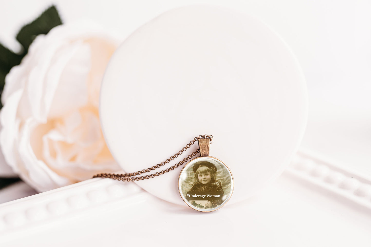 A round pendant necklace on a delicate chain, featuring a vintage-style printed portrait of a young girl with the text 'Underage Woman'. The pendant is displayed on a white circular stand next to a soft white rose.