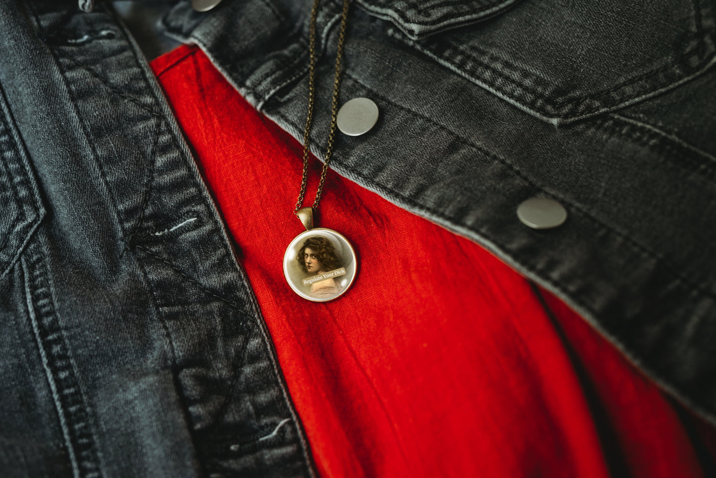 Product photo of a 1-inch glass cabochon pendant necklace featuring “Regulate Your Dick” with a sepia portrait of a curly-haired vintage woman on a 20-inch chain styled over a red shirt and black denim jacket.