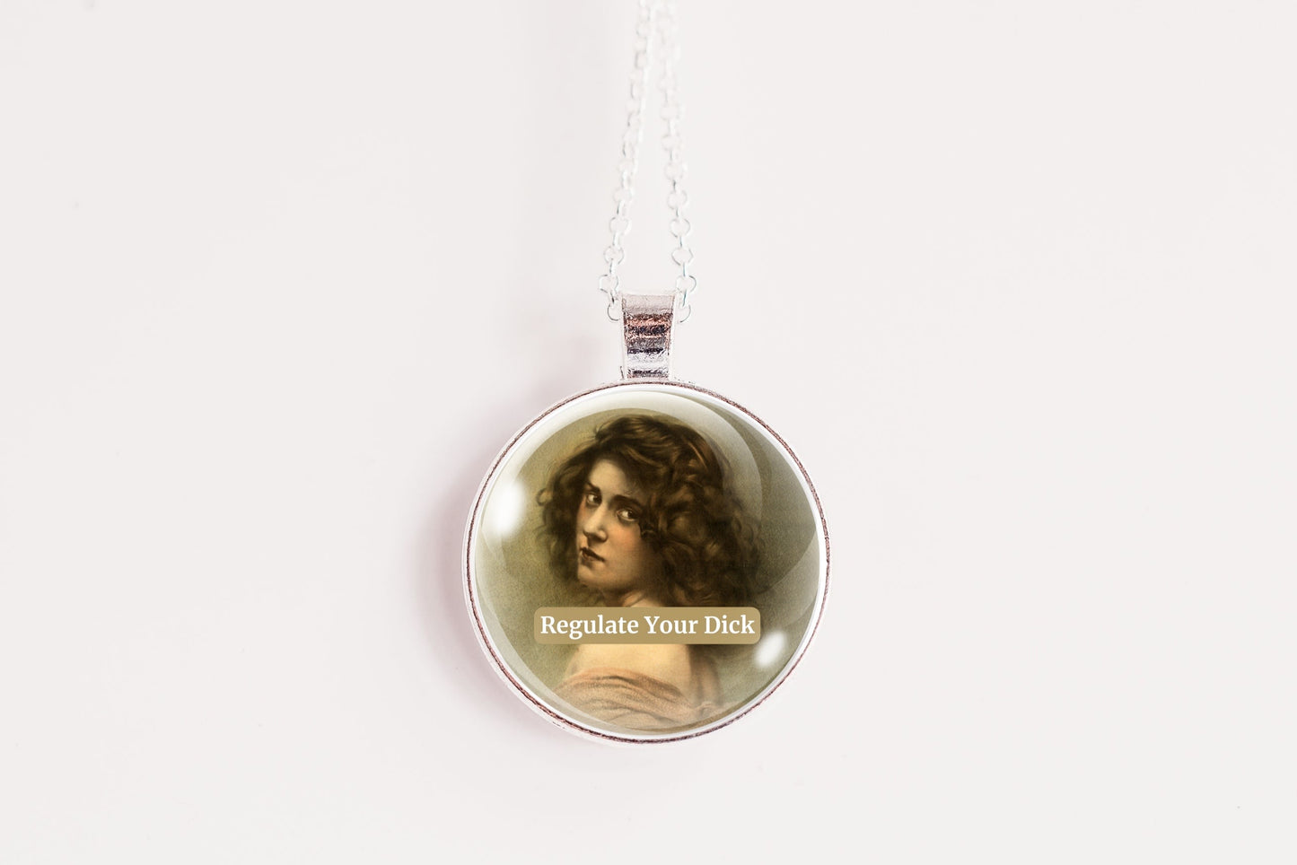 Product photo of a 1-inch glass cabochon pendant necklace featuring “Regulate Your Dick” with a sepia portrait of a curly-haired vintage woman on a 20-inch chain against a plain white background.
