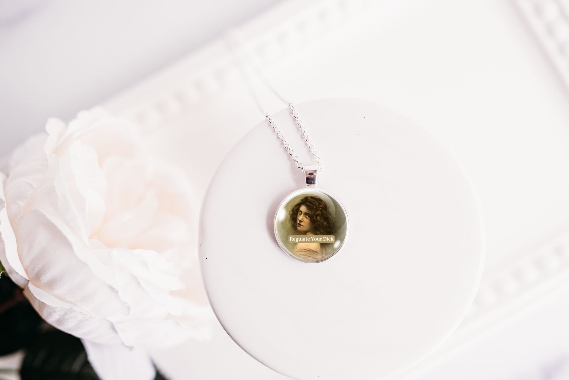 A silver necklace with a round pendant featuring a vintage-style portrait of a woman, displayed on a white circular stand next to a white rose.