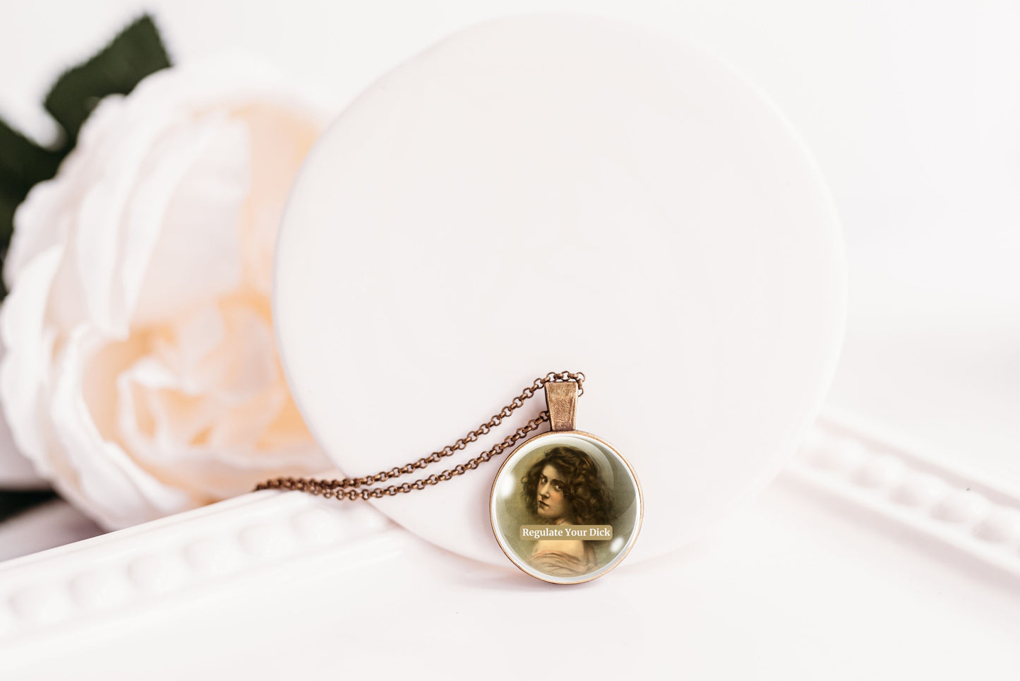 Product photo of a 1-inch glass cabochon pendant necklace featuring “Regulate Your Dick” with a sepia portrait of a curly-haired vintage woman on a 20-inch chain.