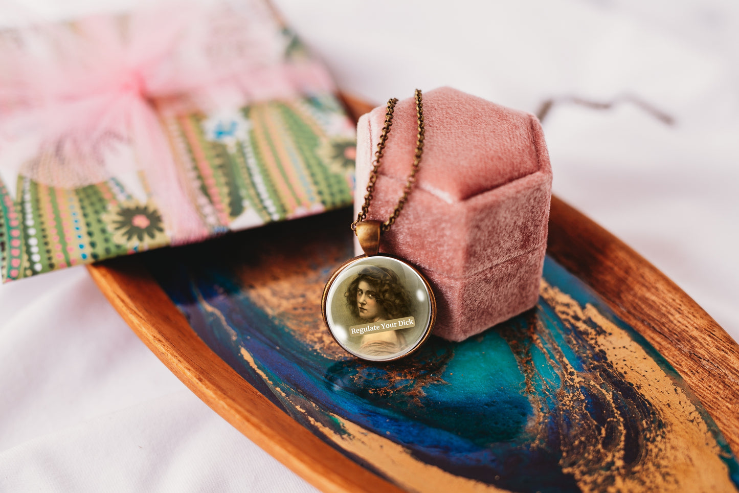 A vintage-style portrait pendant necklace on a bronze chain, displayed on a pink velvet jewelry box resting on a decorative blue and gold tray.