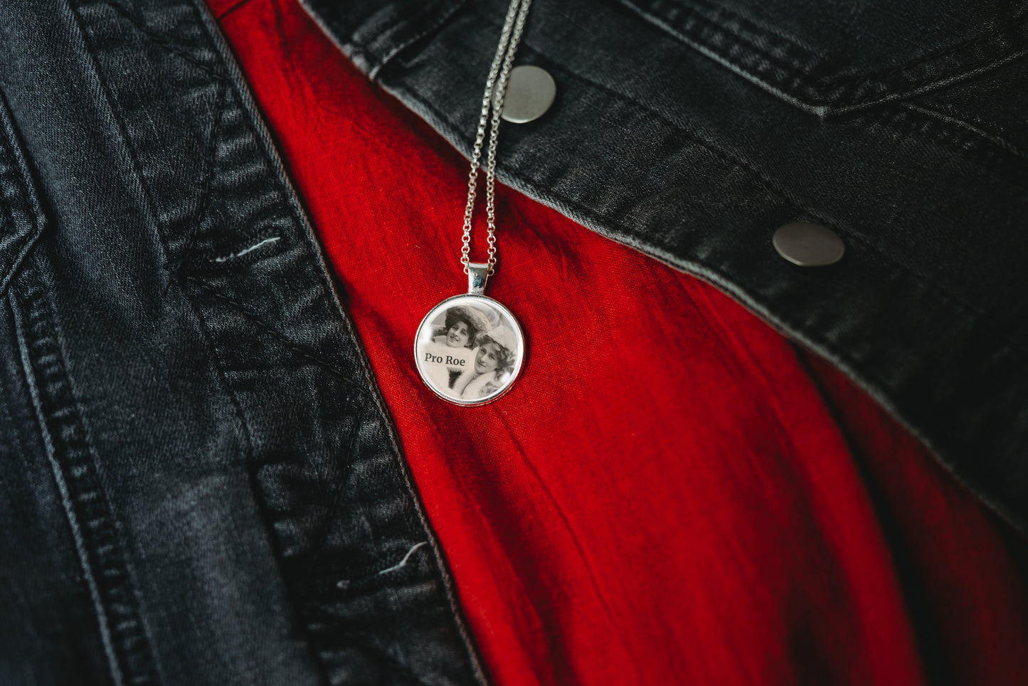Product photo of a 1-inch glass cabochon pendant necklace featuring “Pro Roe” with two vintage women wearing decorative hats on a 20-inch chain, draped over a black denim jacket and a red shirt.
