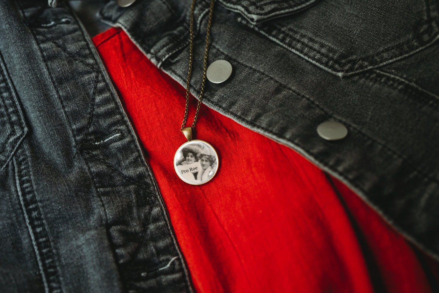 Product photo of a 1-inch glass cabochon pendant necklace featuring “Pro Roe” with two vintage women wearing decorative hats on a 20-inch chain, hanging from a dark chain over a bright red shirt and black denim jacket.