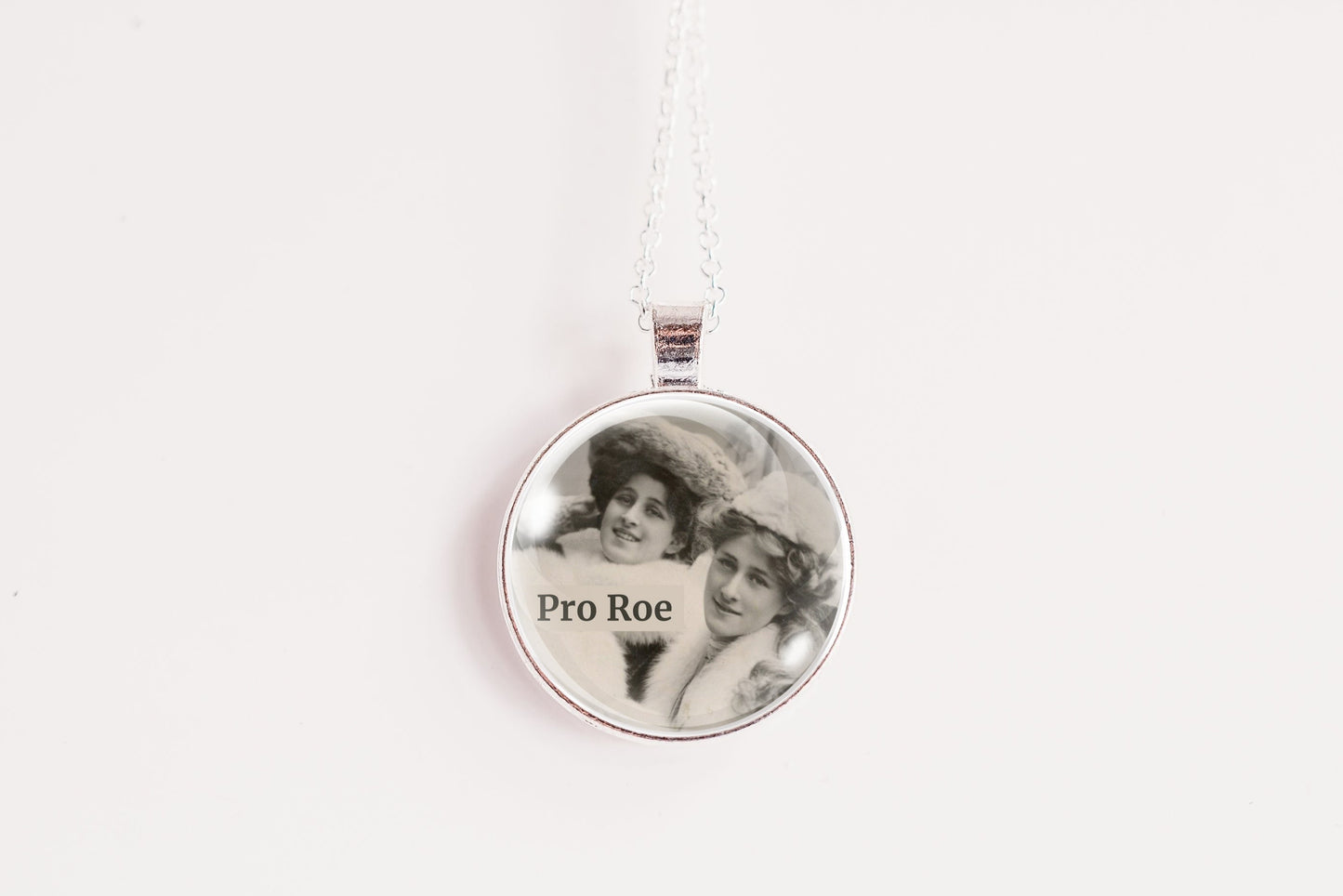 A silver round pendant necklace on a white chain, featuring a custom black-and-white photo of two women with the text 'Pro Roe' printed on the front, set against a plain white background.