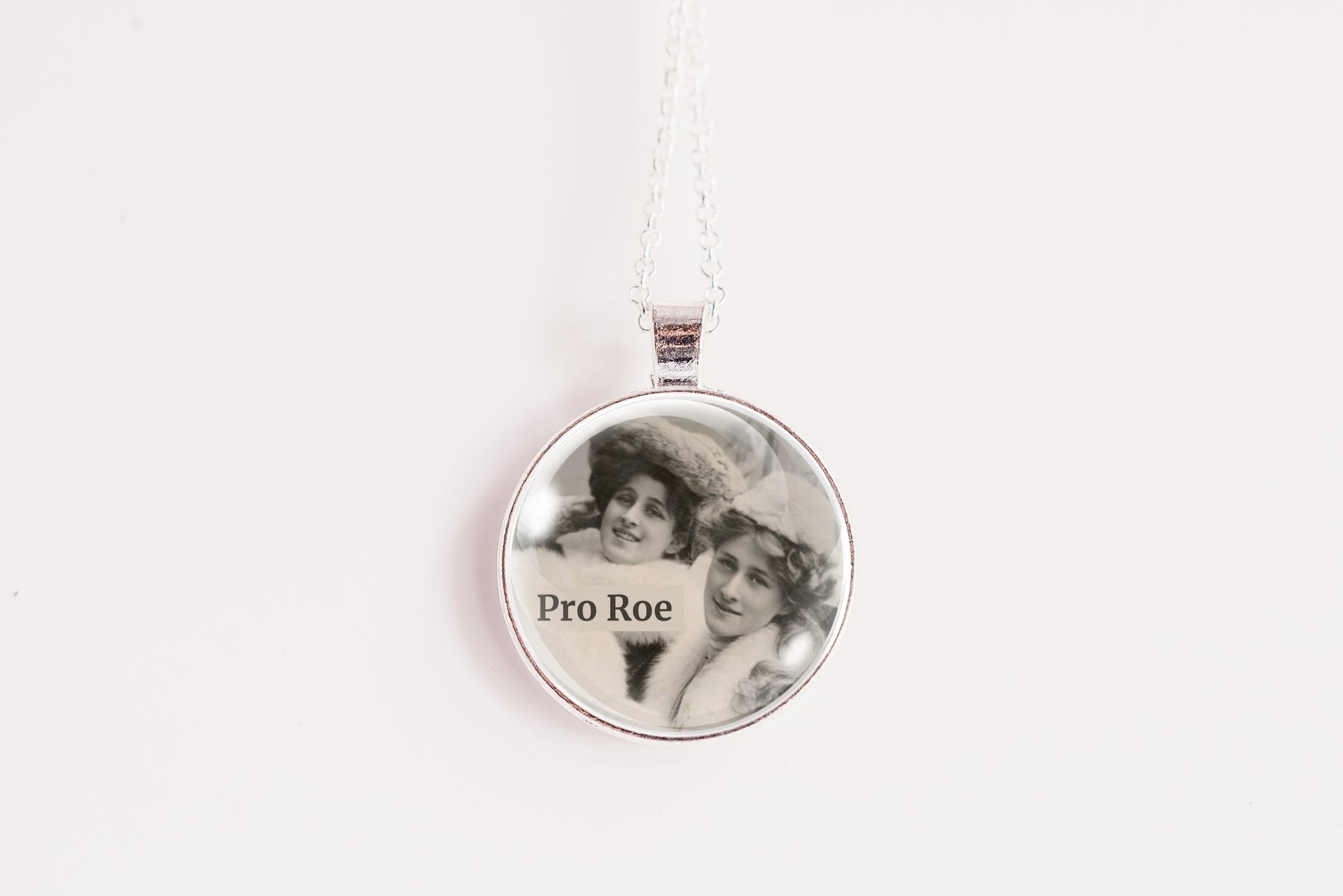 A silver round pendant necklace on a white chain, featuring a custom black-and-white photo of two women with the text 'Pro Roe' printed on the front, set against a plain white background.