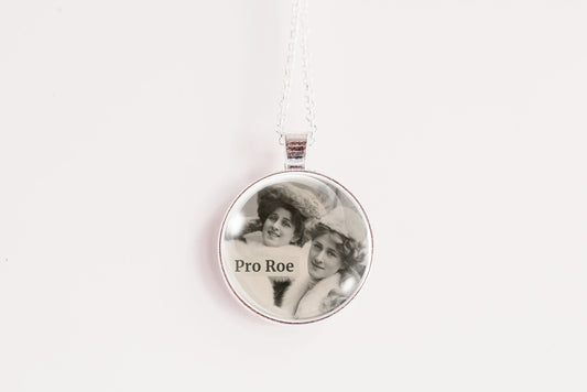 A silver round pendant necklace on a white chain, featuring a custom black-and-white photo of two women with the text 'Pro Roe' printed on the front, set against a plain white background.