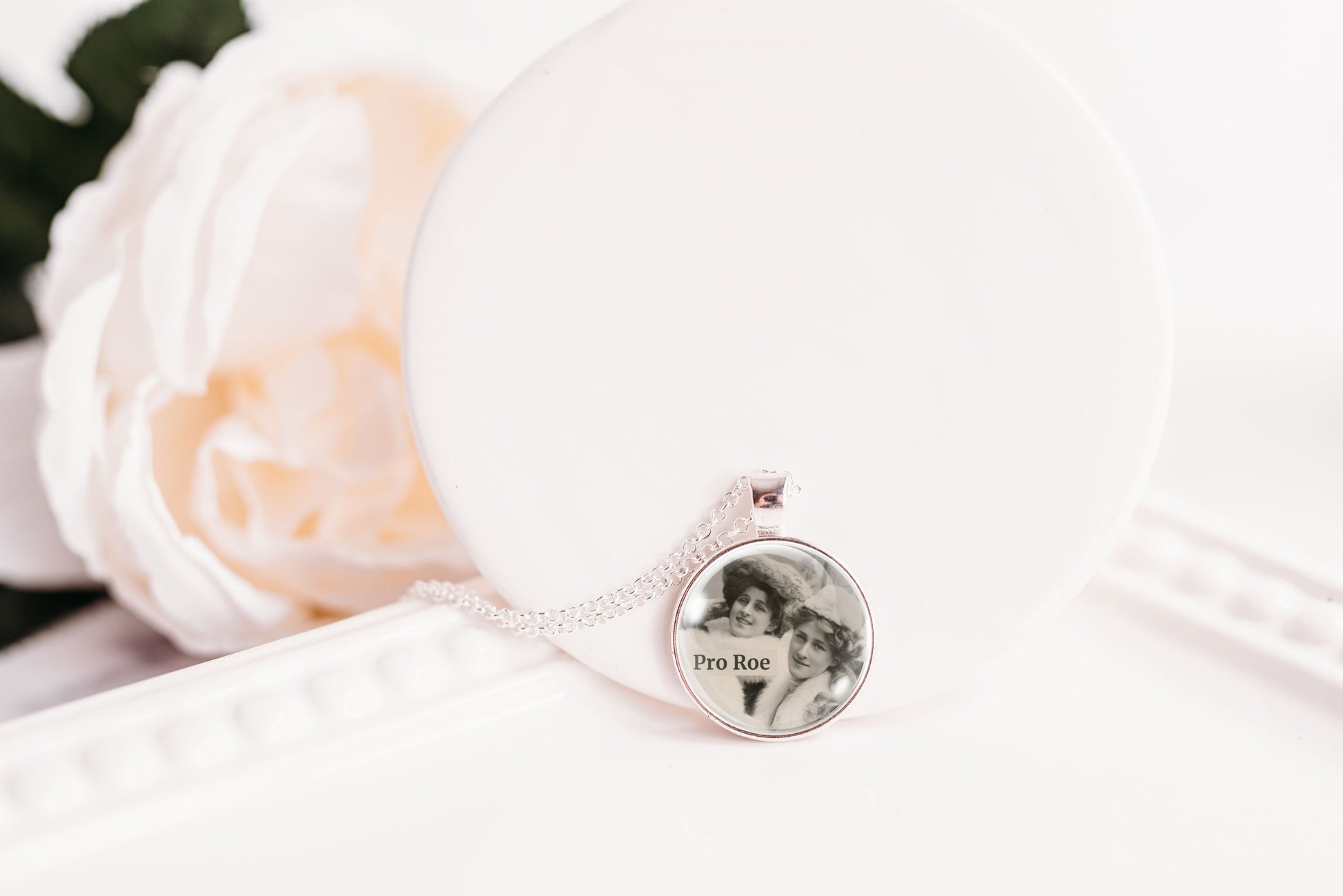 A silver-toned necklace with a round pendant displaying a black-and-white portrait photo, resting on a white display stand next to a soft white rose.