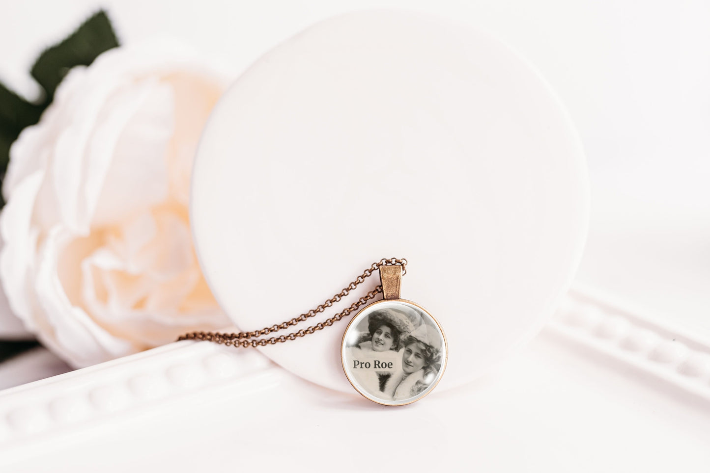 A close-up of a round pendant necklace on a delicate chain, featuring a black-and-white portrait of two women with the text 'Pro Roe' printed inside. The pendant is displayed on a white circular stand next to a soft white rose.