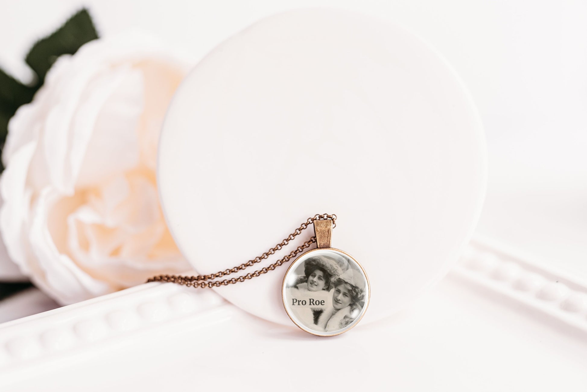 A close-up of a round pendant necklace on a delicate chain, featuring a black-and-white portrait of two women with the text 'Pro Roe' printed inside. The pendant is displayed on a white circular stand next to a soft white rose.