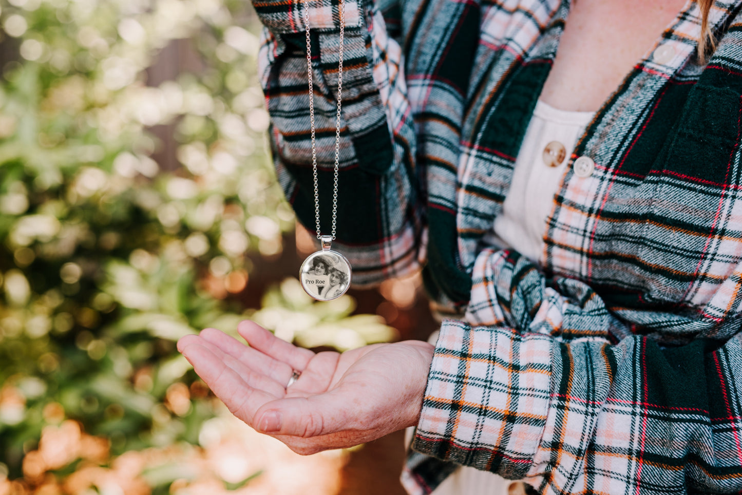 Person wearing a plaid shirt holding a necklace with a pendant outdoors.