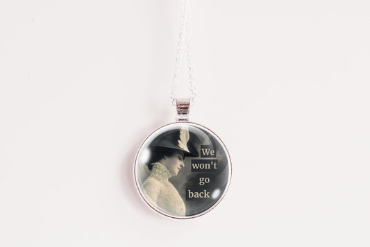 A round pendant necklace hanging from a white cord against a plain white background. The pendant has a silver-toned metal border and a printed design showing a woman in profile on a dark background with the text 'We won't go back' in light lettering.