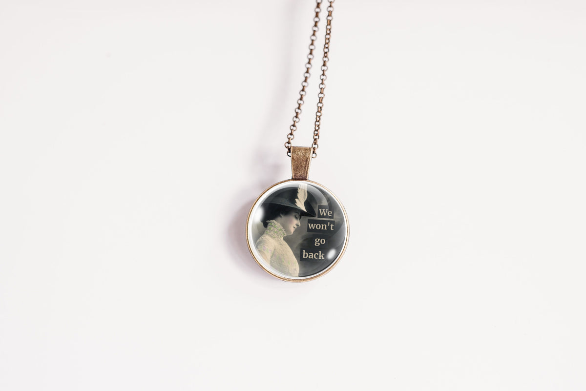 A round pendant necklace on a delicate chain, featuring a printed black-and-white portrait of a woman in a hat with the text 'We won't go back' on a plain white background.