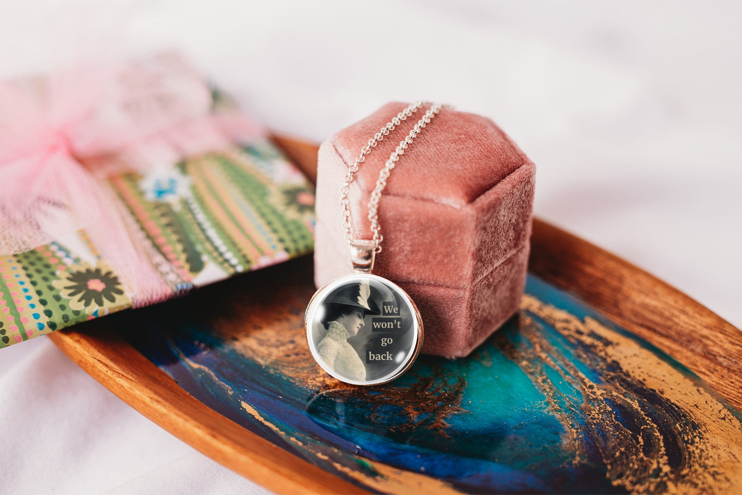 A silver-toned round pendant necklace with text rests on a dusty pink velvet jewelry box, which sits on a decorative wooden tray. 
