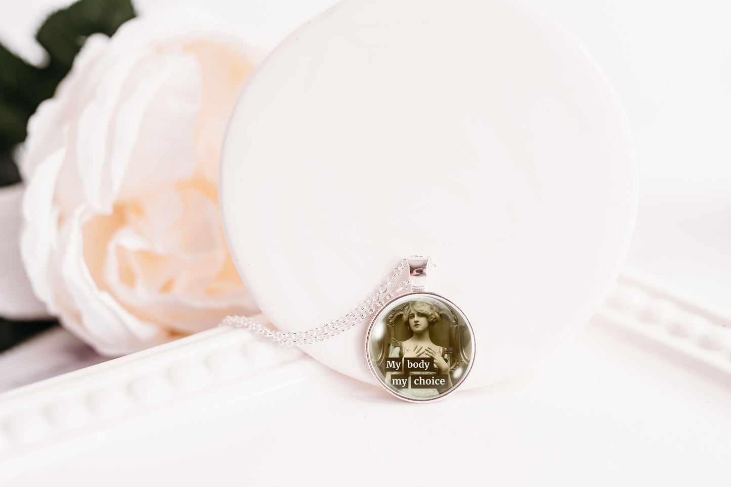 A silver-toned pendant necklace with a circular charm featuring a sepia-toned portrait and the text 'My body my choice', displayed on a white surface next to a white rose.
