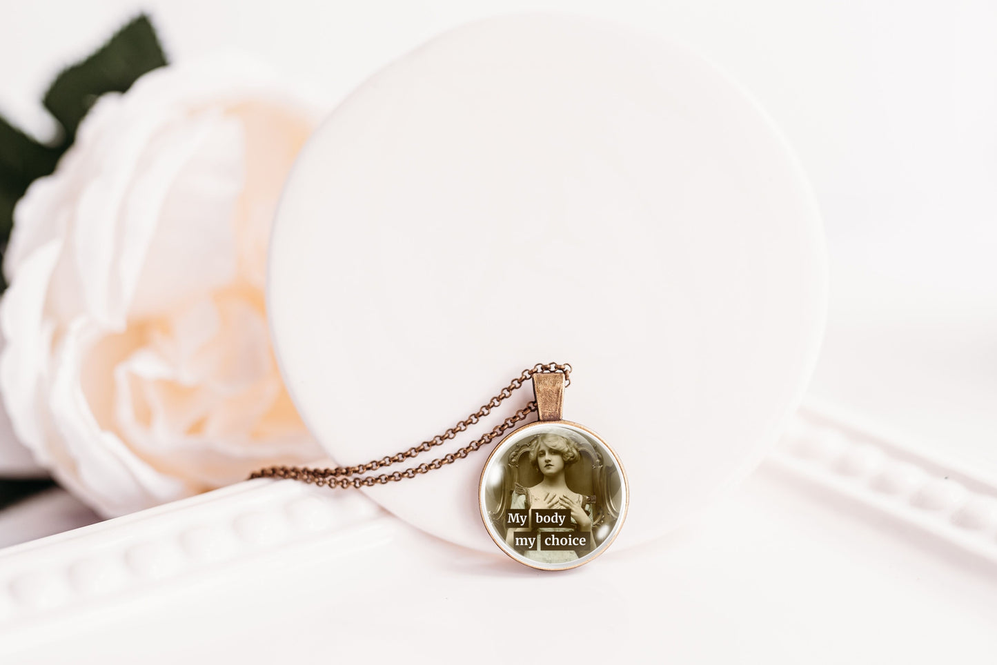 A bronze-toned chain necklace with a round pendant featuring a vintage-style portrait of a woman and the text 'My body my choice' in white lettering, displayed on a white surface next to a soft white flower.