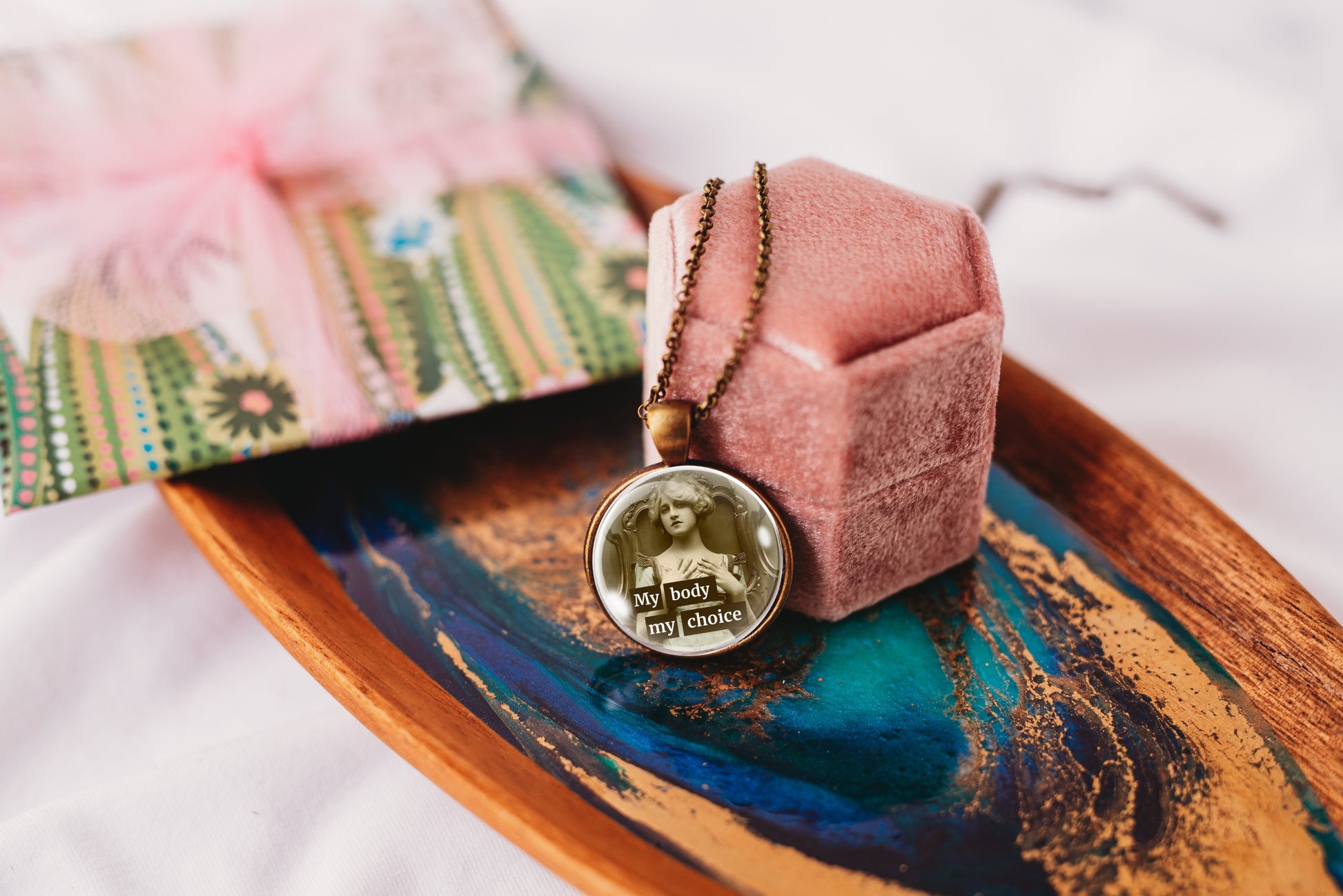 A round pendant necklace with a vintage-style portrait and the text 'My body my choice' resting on a pink velvet jewelry box, placed on a decorative wooden tray with blue and gold accents.