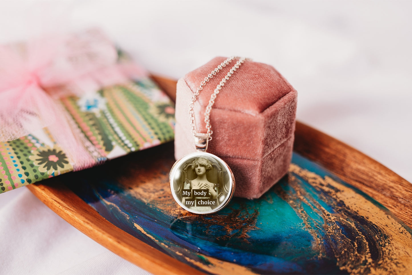A silver-toned round pendant necklace with a photo insert and the text 'My body my choice' rests on a dusty pink velvet jewelry box, which is placed on a decorative wooden tray.