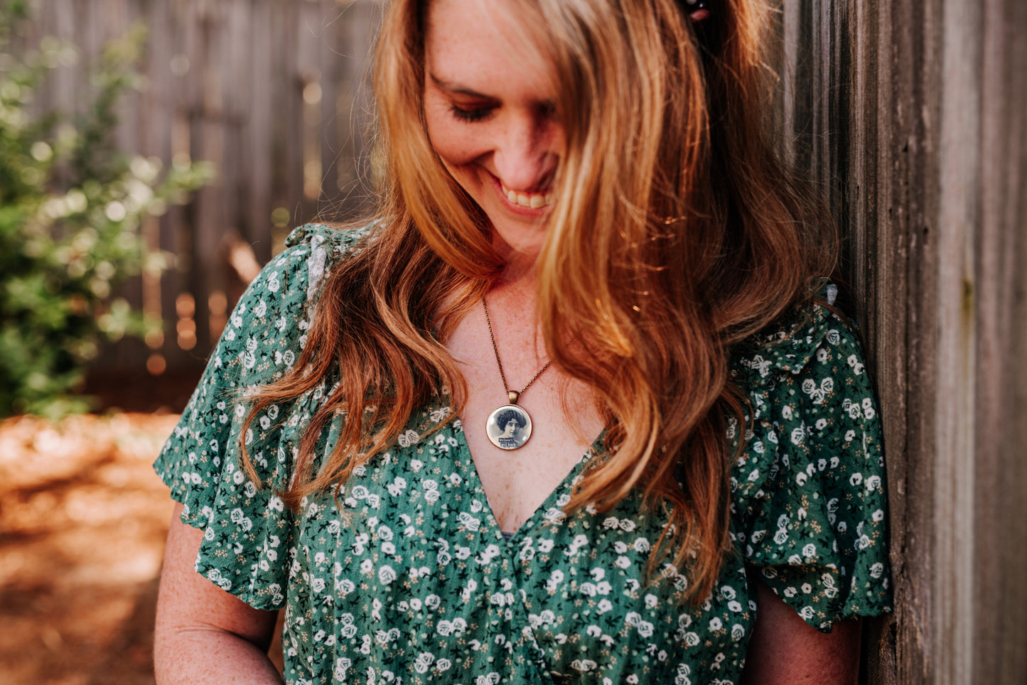 A woman with long wavy light brown hair leans against a weathered wooden fence, wearing a green short-sleeve V-neck dress with a white floral pattern and a silver pendant necklace.