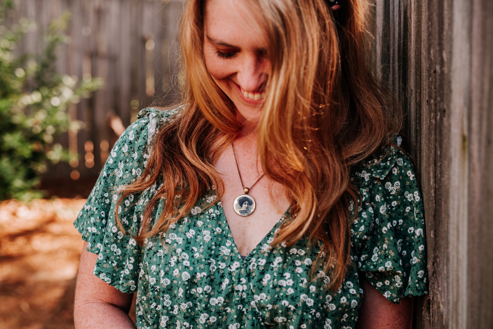 A woman with long wavy light brown hair leans against a weathered wooden fence, wearing a green short-sleeve V-neck dress with a white floral pattern and a silver pendant necklace.