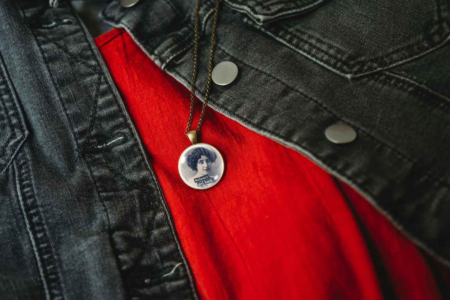 Product photo of a 1-inch glass cabochon pendant necklace featuring “We Won’t Go Back” with a black-and-white portrait of a curly-haired vintage woman on a 20-inch chain over a bright red shirt and black denim jacket.