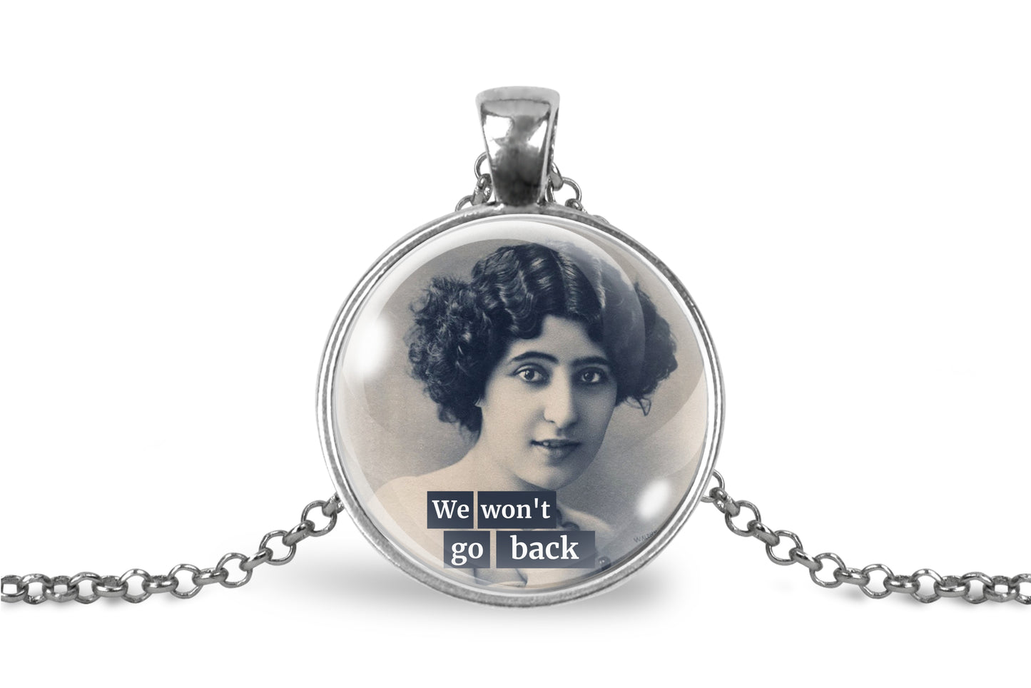 A silver pendant necklace featuring a circular glass dome with a vintage black-and-white portrait of a woman and the text 'We won't go back' in white lettering on dark blocks. The pendant is suspended from a silver chain on a plain white background.