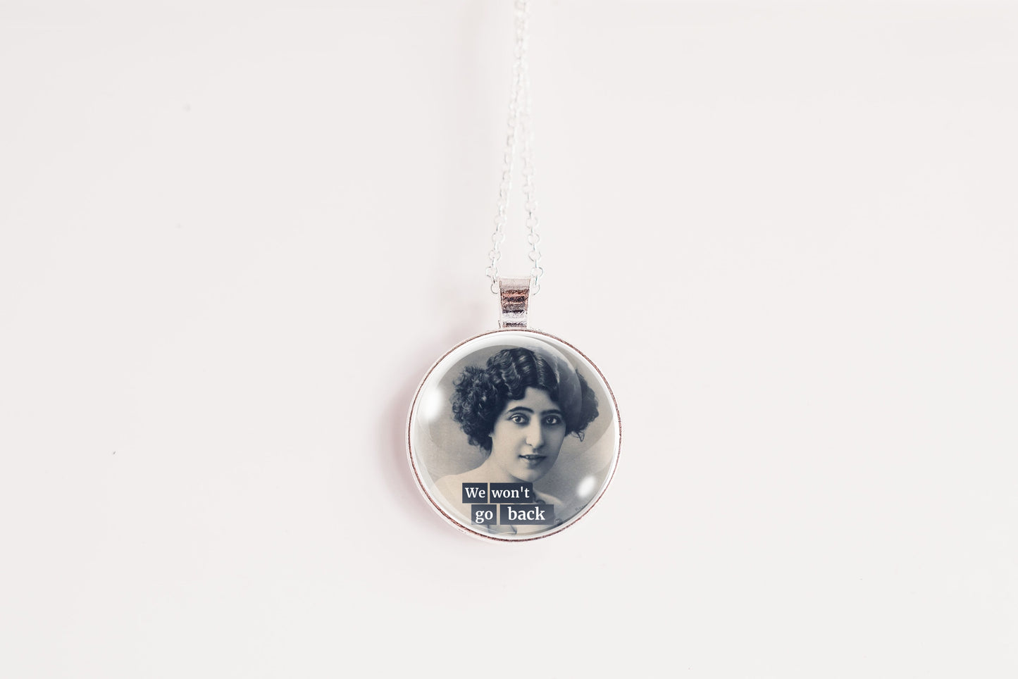 A silver circular pendant necklace featuring a vintage black-and-white portrait of a woman with the text 'We won't go back' at the bottom, suspended on a silver chain against a plain white background.