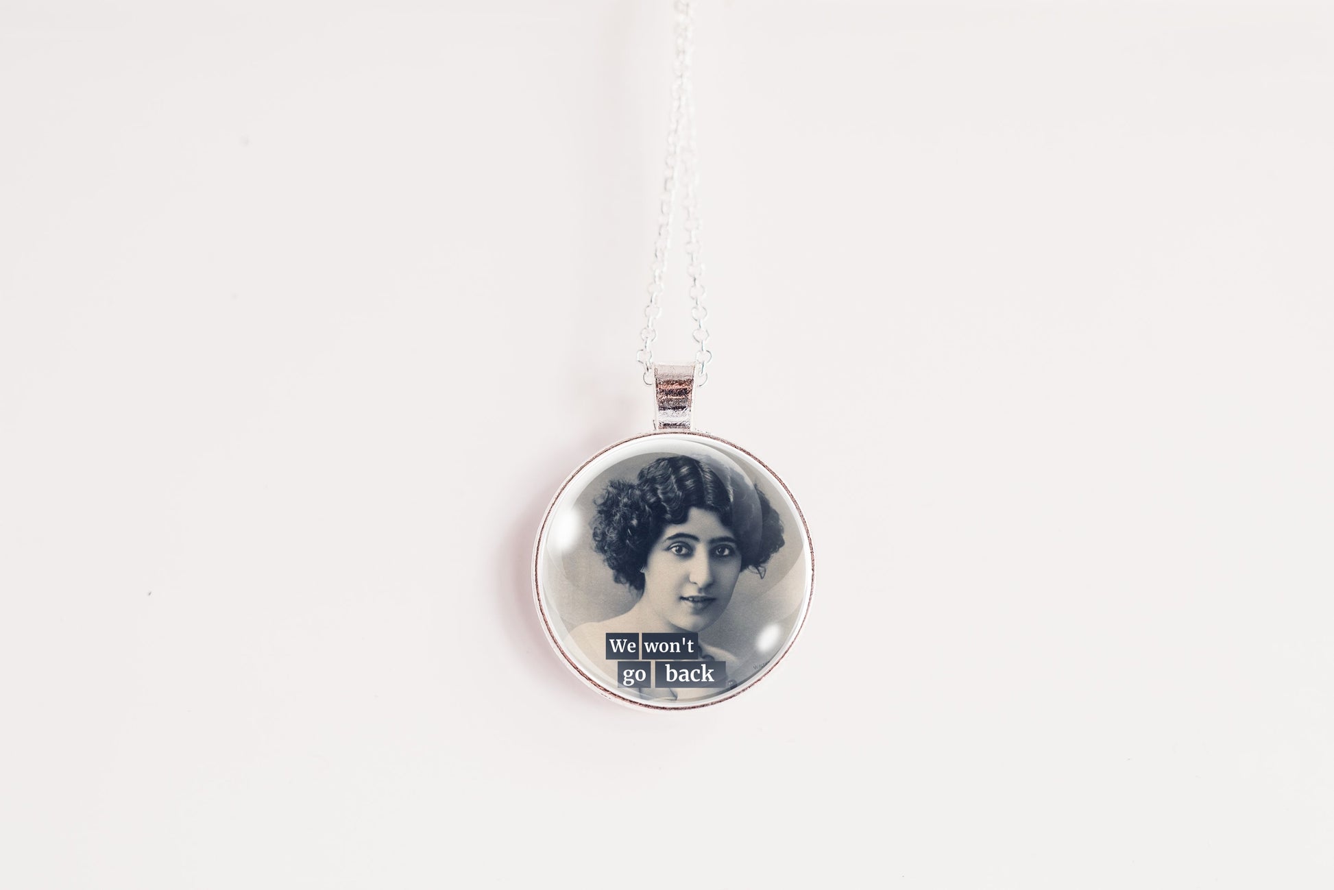A silver circular pendant necklace featuring a vintage black-and-white portrait of a woman with the text 'We won't go back' at the bottom, suspended on a silver chain against a plain white background.