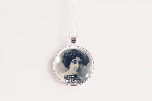 A silver circular pendant necklace featuring a vintage black-and-white portrait of a woman with the text 'We won't go back' at the bottom, suspended on a silver chain against a plain white background.