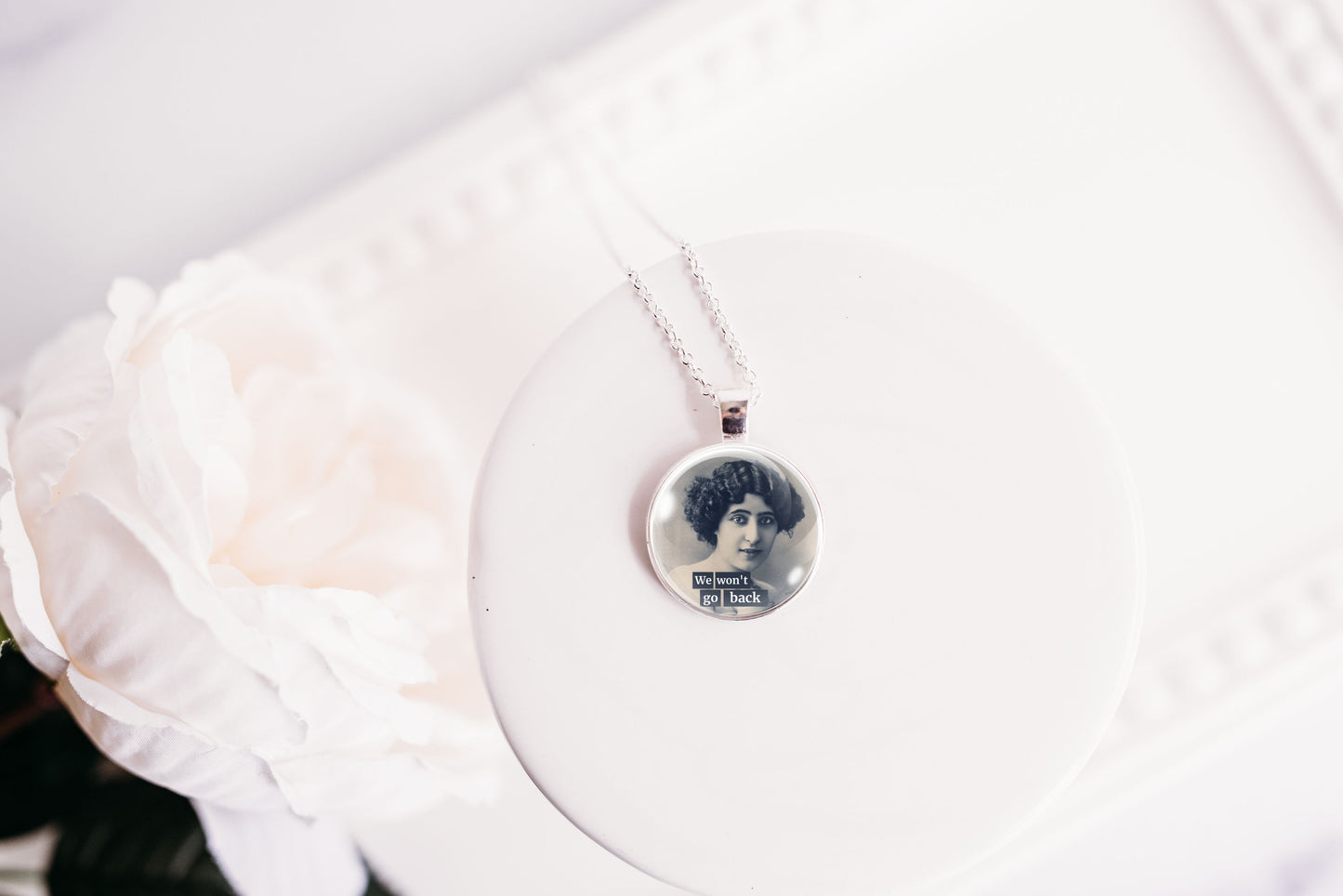 A silver necklace with a round photo pendant displayed on a white circular stand next to a white rose.
