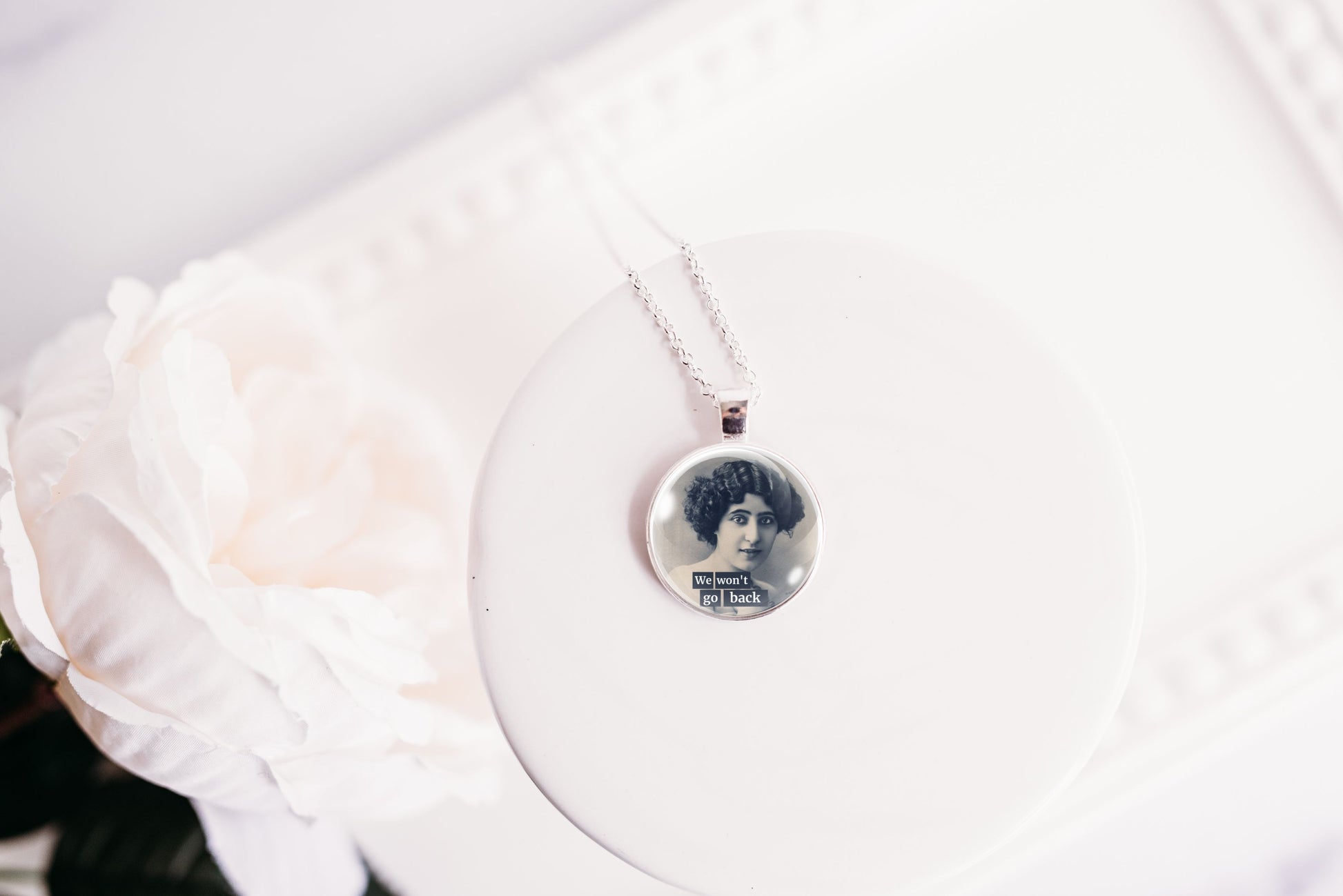 A silver necklace with a round photo pendant displayed on a white circular stand next to a white rose.