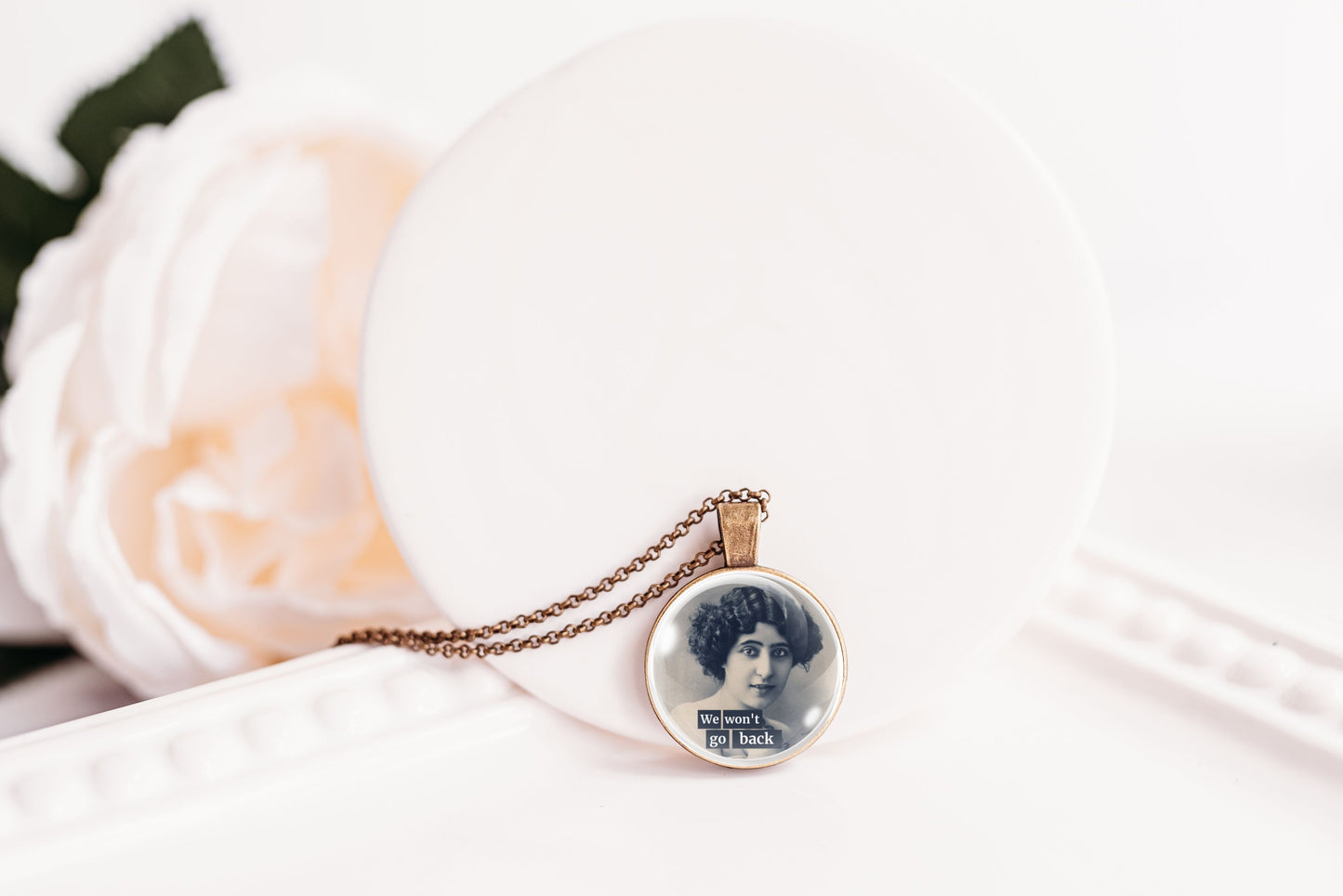 A round pendant necklace with a vintage portrait print and text, hanging from a delicate chain on a white display stand with a soft white flower in the background.