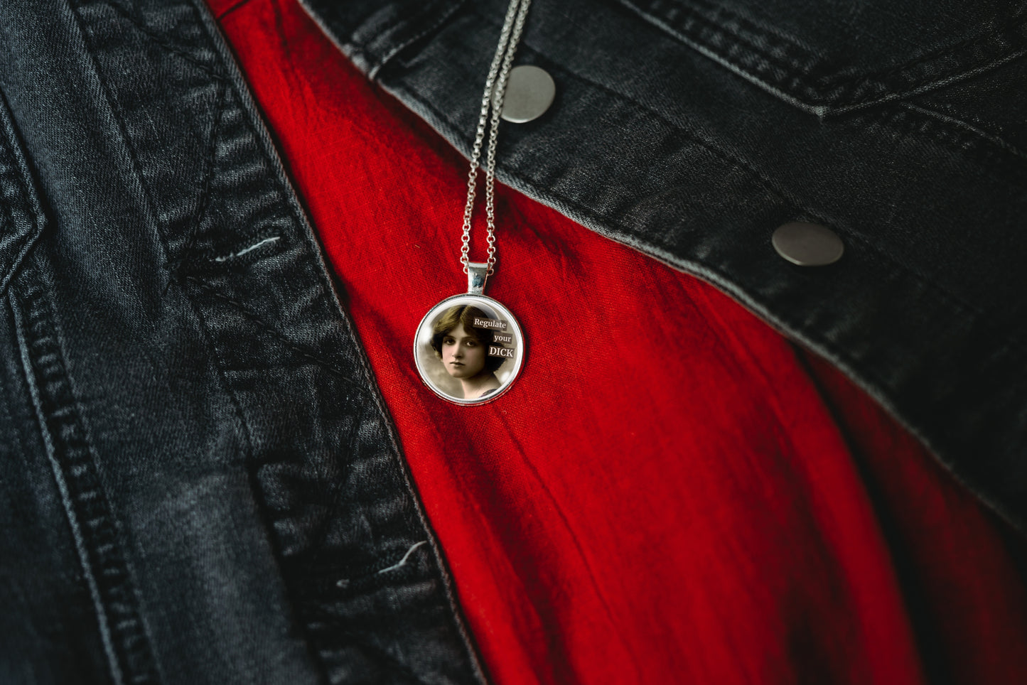 Product photo of a 1-inch glass cabochon pendant necklace featuring “Regulate Your Dick” with a sepia portrait of a vintage-style woman on a 20-inch chain, displayed on a red shirt and black denim jacket.