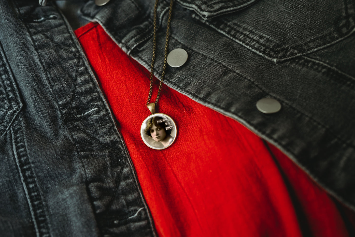Product photo of a 1-inch glass cabochon pendant necklace featuring “Regulate Your Dick” with a sepia portrait of a vintage-style woman on a 20-inch chain, styled over a bright red shirt and a dark gray denim jacket.