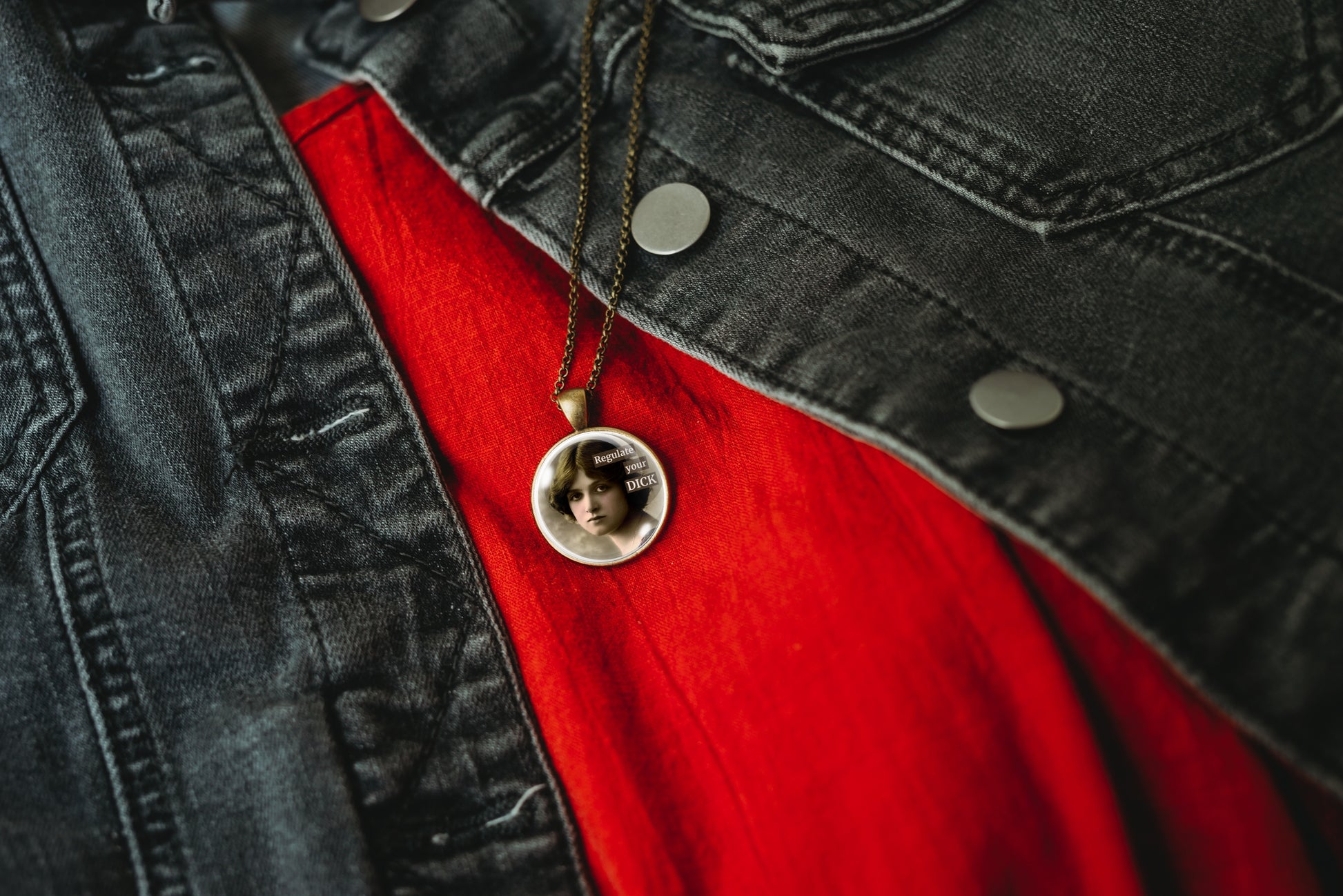 Product photo of a 1-inch glass cabochon pendant necklace featuring “Regulate Your Dick” with a sepia portrait of a vintage-style woman on a 20-inch chain, styled over a bright red shirt and a dark gray denim jacket.