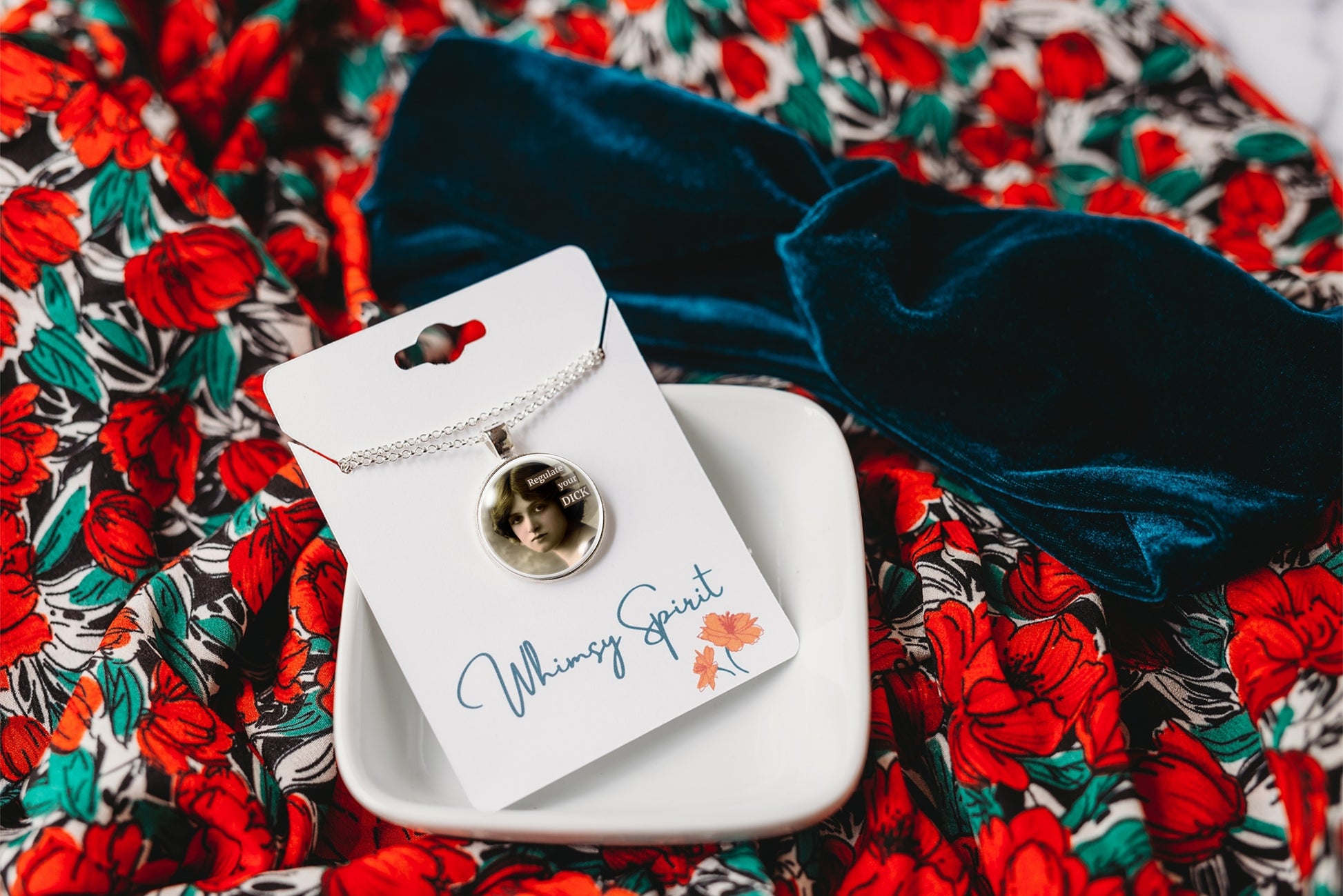 A photo necklace on a delicate chain, displayed on a white card labeled 'Whimsy Spirit, ' resting on a small white dish. The necklace is placed on a dark navy velvet bow over a red and green floral patterned fabric background.