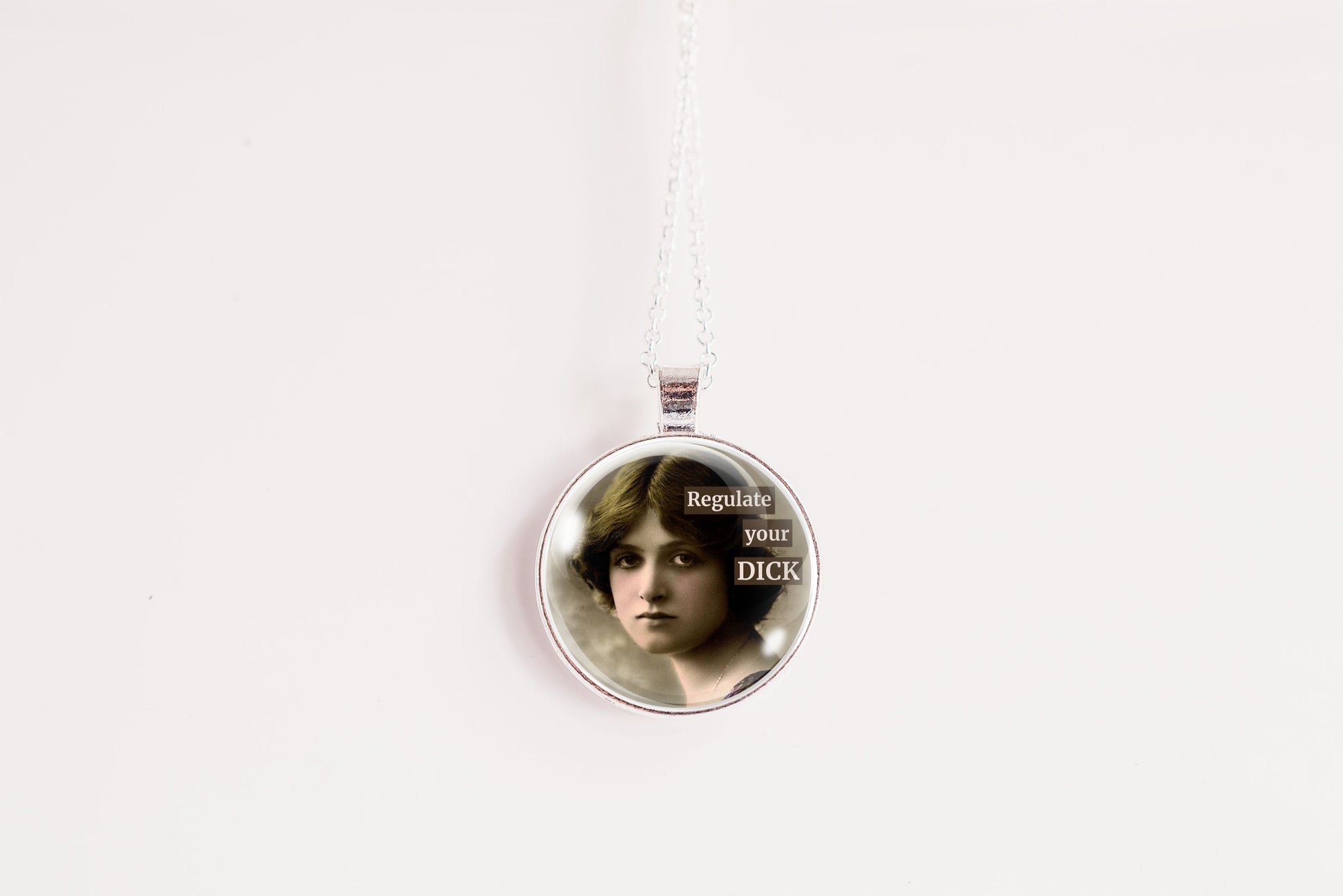 A silver round pendant necklace hanging from a silver chain against a plain white background. The pendant features a printed portrait of a woman with the text 'Regulate your DICK' in white lettering.