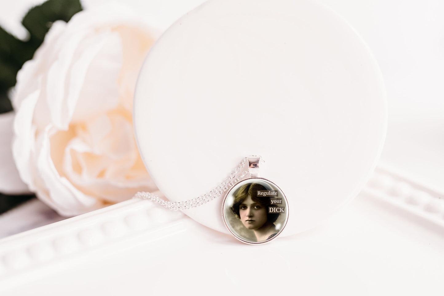 A silver-toned necklace with a round pendant displaying a black-and-white vintage portrait of a woman, resting on a white display stand next to a soft white rose.