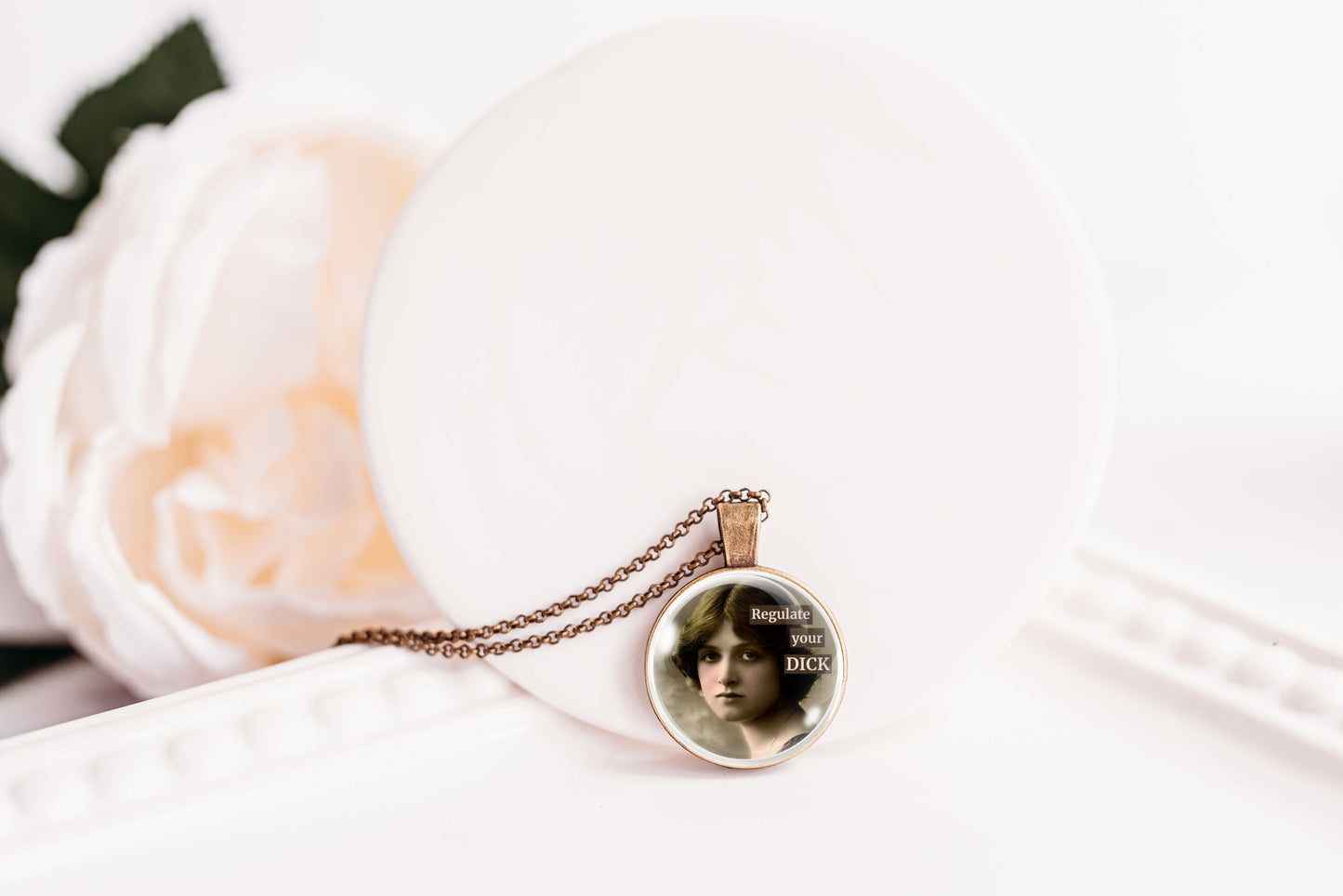 A round pendant necklace with a vintage-style portrait of a woman and the text 'Regulate Your DICK' on a delicate chain, displayed on a white circular stand with a soft-focus white flower in the background.