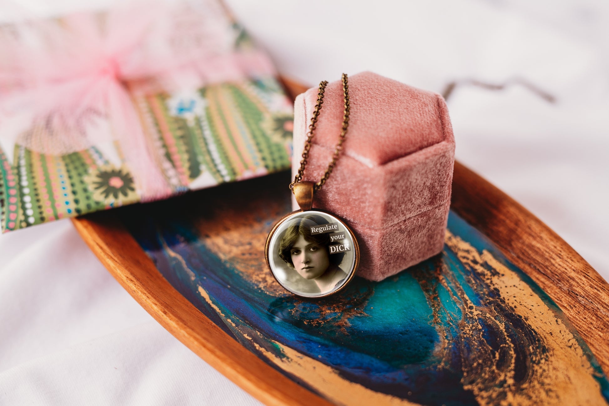 Product photo of a 1-inch glass cabochon pendant necklace featuring “Regulate Your Dick” with a sepia portrait of a vintage-style woman on a 20-inch chain displayed on a pink velvet ring box, resting on a decorative wooden tray 