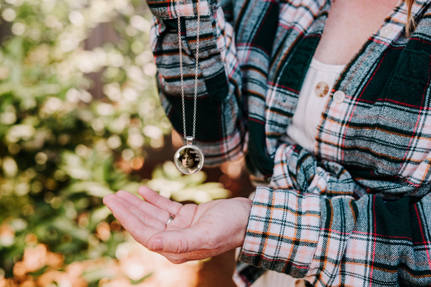 A close-up lifestyle shot of a person wearing a green, black, and white plaid flannel shirt, holding their open hand out to display a silver-toned pendant necklace with a circular portrait 