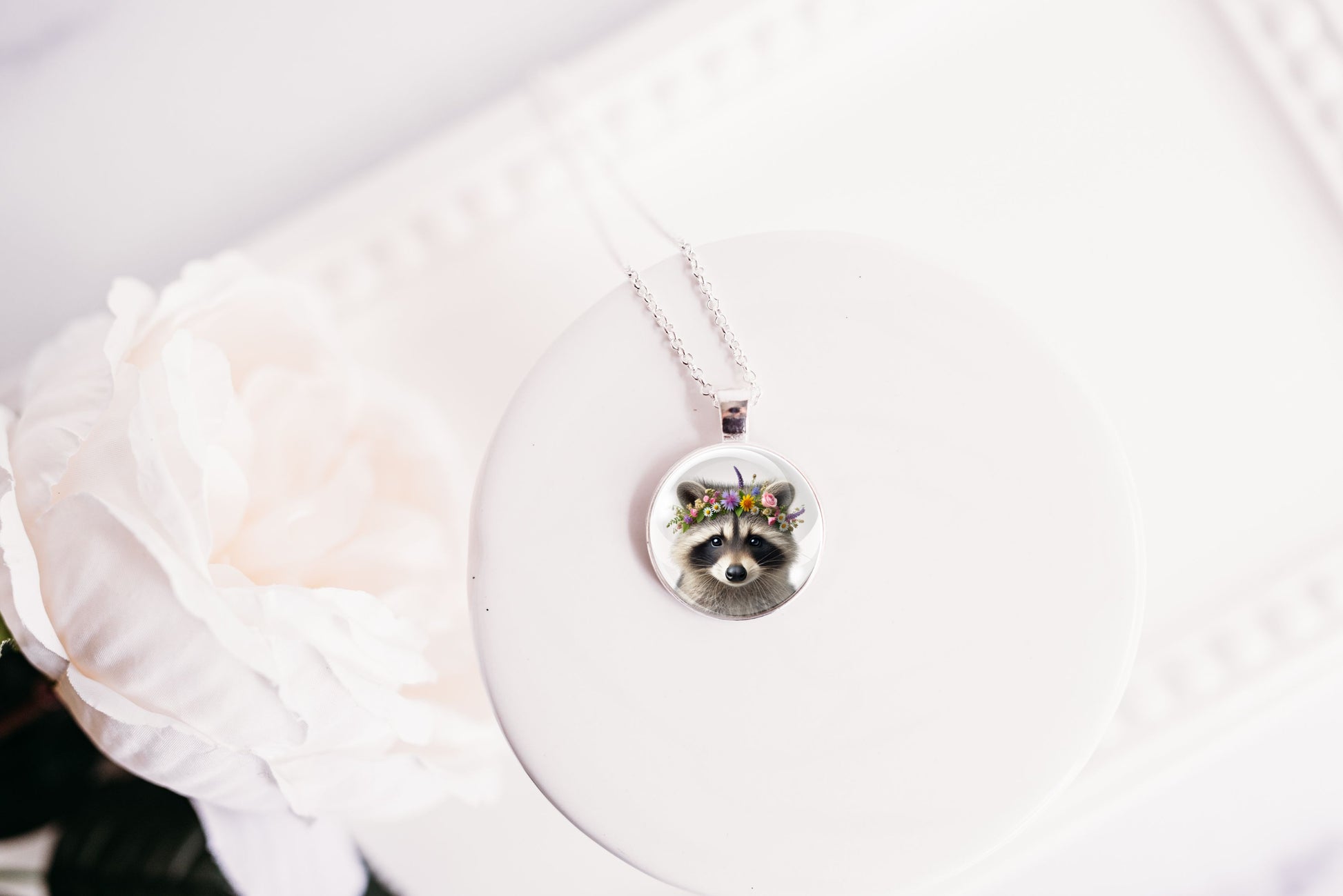Close up image of a raccoon wearing a whimsical flower crown set in a silver-toned bezel. 