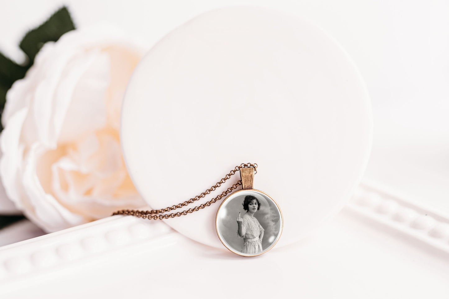 A round pendant necklace with a black-and-white portrait photo in the center, hanging from a bronze-toned chain. The pendant is displayed on a white circular stand next to a soft white rose.