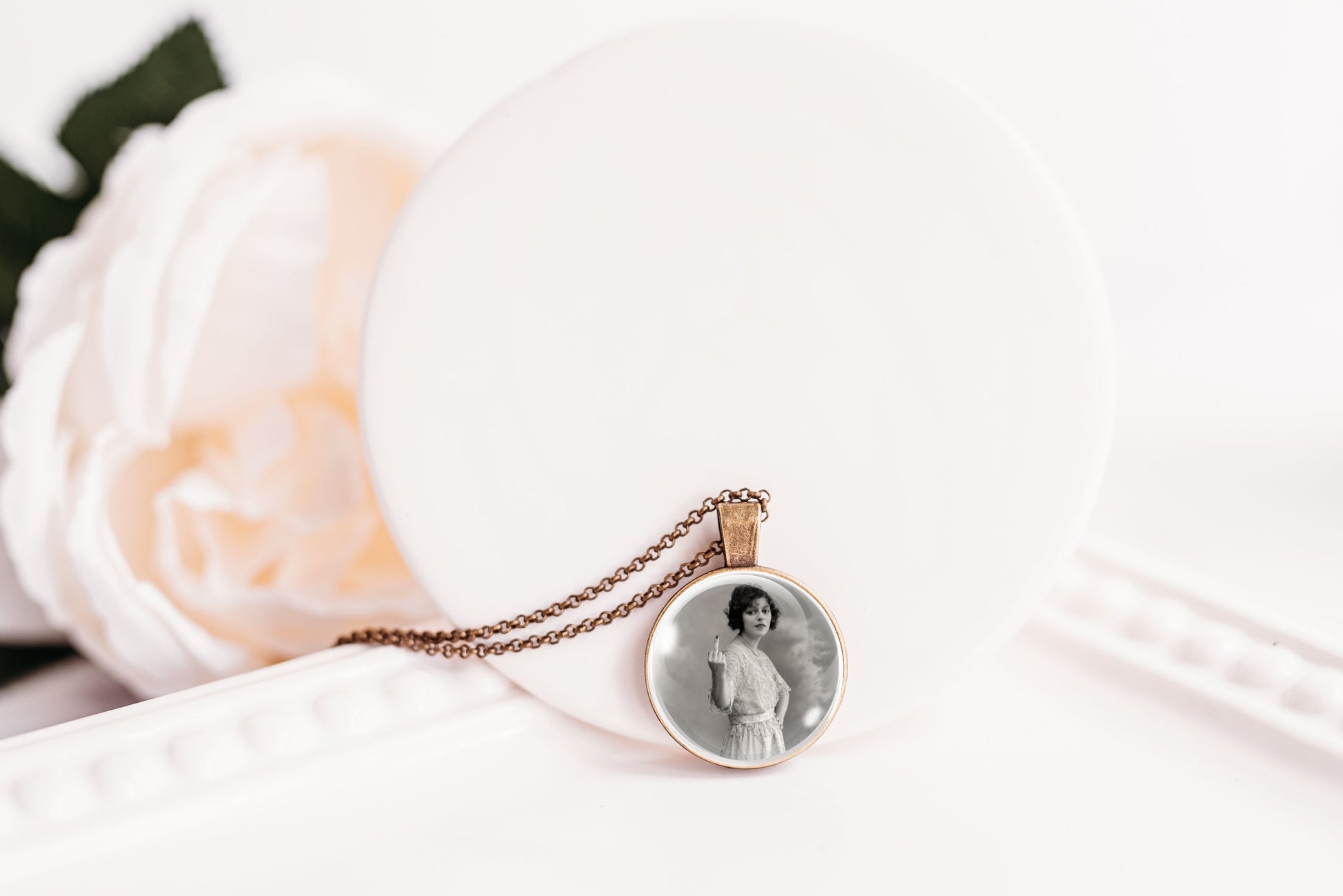 A round pendant necklace with a black-and-white portrait photo in the center, hanging from a bronze-toned chain. The pendant is displayed on a white circular stand next to a soft white rose.